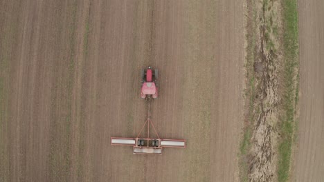 Overhead,-look-down-view-from-drone-tracking-large-red-farm-tractor-towing-machinery-as-farmer-works-in-field-to-prepare-land-for-planting
