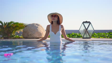 A-young-woman-stands-in-the-water-of-a-resort-pool-wearing-a-bathing-suit,-sunglasses,-and-straw-sun-hat