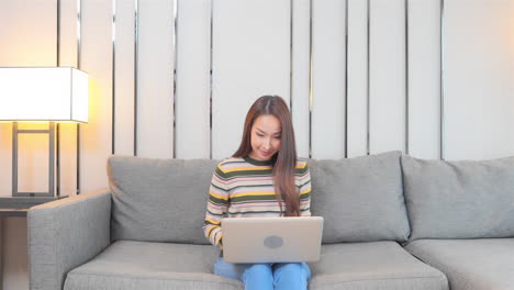 A-young-woman-sitting-on-a-couch-works-on-her-laptop-suddenly-reacts-to-her-screen-by-raising-her-arms-in-success