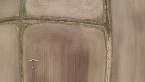 Aerial-look-down-view-from-drone-hovering-over-brown-farm-field-as-tractor-tows-piece-of-equipment-across-soil-below