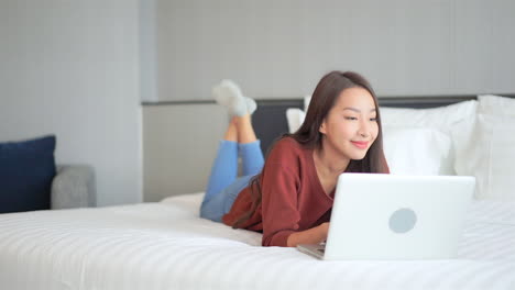 A-young-woman-works-on-her-laptop-as-she-lays-on-her-stomach-on-a-big-comfortable-hotel-bed