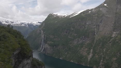 Forward-aerial-reveal-shot-of-Geiranger-fjord-covered-in-snow-on-a-cloudy-day,-Norway