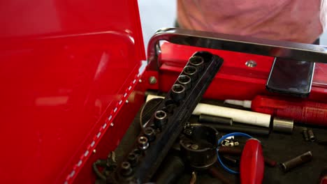 Mechanic-white-caucasian-man-sets-his-cell-phone-android-down-on-red-toolbox,-grabs-socket-wrench-to-work-on-car,-as-camera-pans-down-to-reveal-toolbox-with-tools-and-equipment