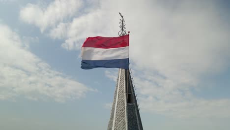 Bandera-Holandesa-Roja,-Blanca-Y-Azul-En-La-Torre-De-La-Iglesia-Puntiaguda-Con-Nubes-Detrás