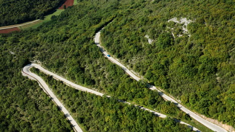 Vista-Aérea-De-Un-Coche-Azul-Conduciendo-Por-Una-Sinuosa-Carretera-De-Montaña-En-Istria,-Croacia