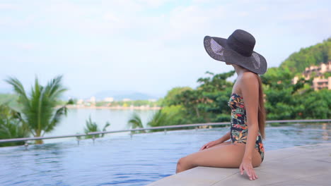 A-pretty-woman-in-a-bathing-suit-and-floppy-sun-hat-sits-on-the-edge-of-a-swimming-pool-with-the-ocean-and-city-skyline-in-the-distance