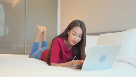 Woman-lying-on-the-bed-with-her-laptop-in-front-of-her-and-typing-message-on-the-keyboard,-happy-smiling-expression-on-her-face,-slow-motion-perspective-view