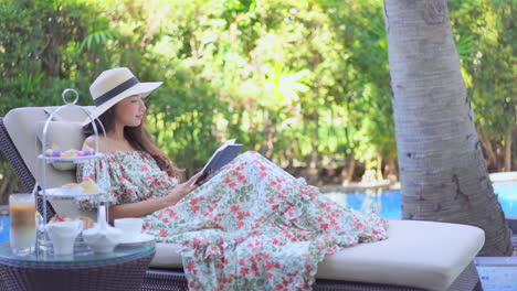 Woman-in-sarafan-sundress-and-hat-lying-on-a-deckchair-and-reading-a-book-outdoors-daytime-in-a-tropical-resort-near-the-swimming-pool