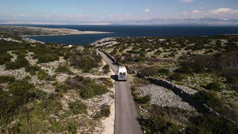 Plain-straight-shot-with-drone-behind-a-camper-van-with-low-altitude-watching-the-sea-on-the-horizon-blue-sky-and-green-in-the-mountains-sunny-day-4k