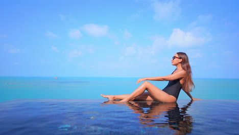 Sexy-young-woman-sunbathing-on-edge-of-infinity-pool-with-blue-ocean-in-background