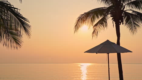 Ein-Einsamer-Sonnenschirm-Sitzt-Zwischen-Zwei-Kokosnusspalmen,-Die-Von-Einem-Leuchtenden-Orange-Und-Gelben-Sonnenuntergang-Unterstützt-Werden