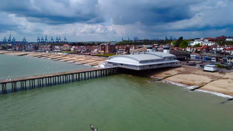 Seagulls-flying-eyelevel-and-under-the-drone-as-it-captures-a-sliding-footage-revealing-the-Felixstowe-Pier-in-Suffolk,-its-town,-shipping-industry,-beachfront,-townhouse,-big-rain-clouds