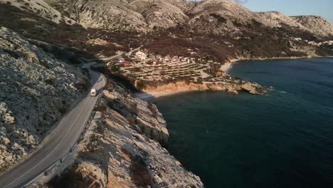 Aerial-of-Camper-driving-on-Krk,-Croatia-along-the-coast-with-a-spectacular-sunset-in-the-Adriatic-Sea,-vanlife-in-a-sunny-day
