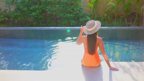 Back-view-of-woman-sitting-on-pool-edge-with-orange-swimsuit-and-hat
