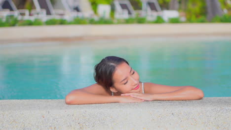 Close-up-of-a-pretty-young-woman-resting-her-head-on-the-arms-along-the-edge-of-a-resort-swimming-pool