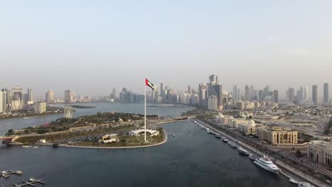 Aerial-view-of-the-Flag-of-the-United-Arab-Emirates-waving-in-the-air-city-development-in-Background,-The-national-symbol-of-UAE-over-Sharjah's-Flag-Island