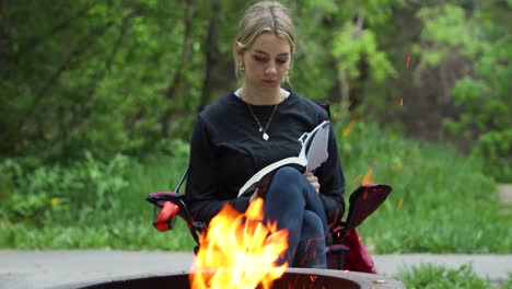 Young-woman-peacefully-reading-book-beside-campfire,-relaxing-outdoors-vacation