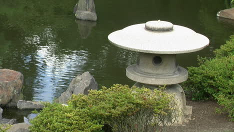 A-Japanese-snow-lantern-stands-amid-azalea-bushes-at-edge-of-pond