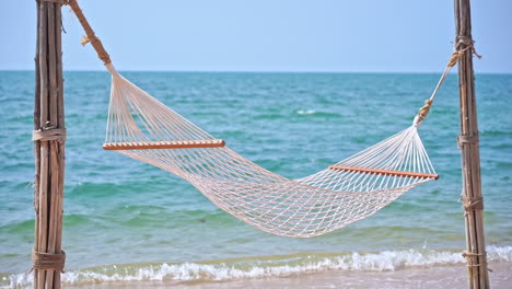 Empty-white-hammock-hangs-from-poles-on-tropical-island-beach-with-calm-turquoise-sea-in-background-and-tiny-waves-lapping-at-the-sandy-beach