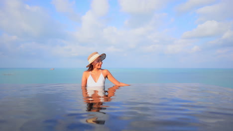 Fittes-Frauenmodell-Im-Weißen-Badeanzug,-Das-Die-Arme-Am-Rand-Des-Infinity-Pools-Auf-Türkisfarbenem-Meereslandschaftshintergrund-In-Bali-An-Einem-Sonnigen-Tag-In-Zeitlupe-Lehnt
