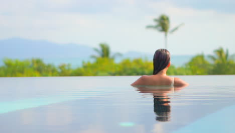 Back-of-sexy-exotic-woman-in-infinity-pool-enjoying-in-water,-view-of-a-tropical-landscape-on-hot-summer-day,-slow-motion-full-frame