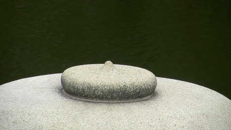 Nahaufnahme-Der-Spitze-Einer-Japanischen-Laterne-Mit-Teichwasser,-Das-Sich-Im-Hintergrund-Bewegt