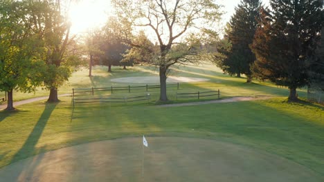 Golf-course-putting-green,-white-pin-flag-waves-in-breeze-during-sunrise
