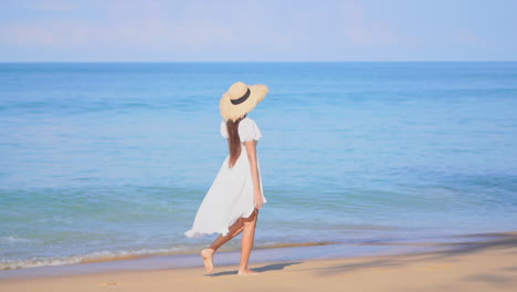 Barefoot-woman-with-white-dress-walking-on-sandy-beach-with-large-straw-hat
