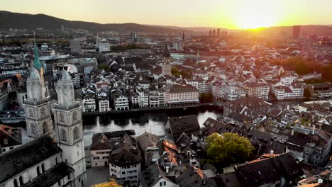 Aerial-flyover-alongside-Limmat-river-in-Zurich,-Switzerland-at-sunset-behind-Grossmünster-church