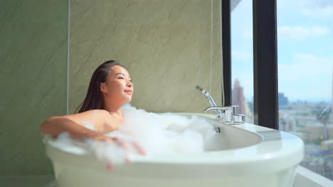 Relaxed-sexy-Asian-woman-taking-bath-in-bathtub-of-luxurious-hotel-bathroom-with-bubbles-and-foam,-cityscape-in-background