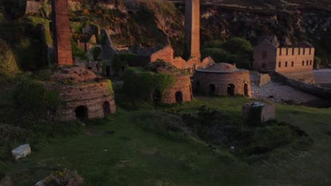 Porth-Wen-abandoned-coastal-derelict-brickworks-remains-golden-sunrise-countryside-bay-aerial-rising-view-pull-back