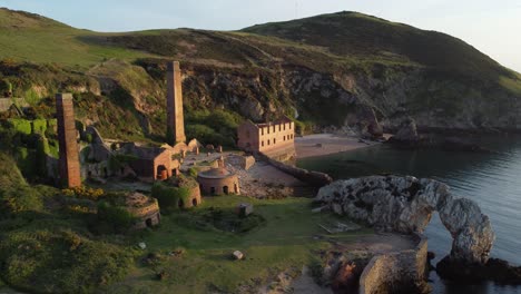 Porth-Wen-abandoned-coastal-derelict-brickworks-remains-golden-sunrise-countryside-bay-aerial-view-wide-orbit-right-across-bay