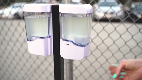 A-woman-using-an-automatic-gel-hand-sanitizer-dispenser-station-outside-at-a-park-in-front-of-a-chainlink-fence-during-the-COVID19-coronavirus-health-pandemic