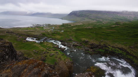Mountain-river-cascading-downslope-towards-bay-in-fjord-Iceland,-pan-left