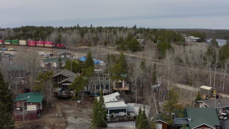 Aerial-ascend-over-ice-and-cottages-to-reveal-a-colorful-freight-train-hauling-large-shipping-containers-through-the-rocky-Canadian-shield