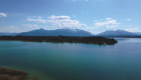 Scenic-4K-aerial-view-upon-Bavaria's-famous-lake-Chiemsee-with-its-castle-island-Herrenchiemsee-and-a-ferry-in-the-rural-countryside-with-a-beautiful-blue-sky-and-the-alps-mountains-in-the-background