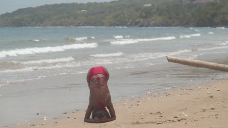Biegsames-Mädchen-In-Einem-Bikini,-Das-Einen-Handstand-Macht,-Während-Die-Wellen-Des-Ozeans-Im-Hintergrund-Zusammenbrechen