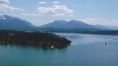 Scenic-aerial-view-upon-Bavaria's-famous-lake-Chiemsee-with-its-castle-island-Herrenchiemsee-and-a-ferry-in-the-rural-countryside-with-a-beautiful-blue-sky-and-the-alps-mountains-in-the-background