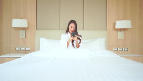 A-young-woman-in-a-bathrobe-is-sitting-up-in-a-luxurious-hotel-bed-texting-from-her-smartphone-to-the-rest-of-the-world
