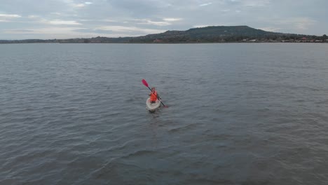 Luftaufnahme,-Die-Einen-Mann-In-Einer-Orangefarbenen-Schwimmweste-Verfolgt,-Der-Am-Frühen-Morgen-Auf-Einem-Kajak-Auf-Dem-Viktoriasee-Paddelt