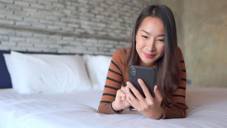 Lächelnde-Asiatische-Frau,-Die-Auf-Dem-Bett-Liegt-Und-Mit-Dem-Finger-Auf-Dem-Handybildschirm-Wischt,-Gesichtsnahaufnahme
