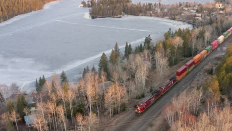 A-long-train-carrying-lots-of-shipping-containers-traveling-through-the-forest-beside-a-frozen-lake-in-Canada