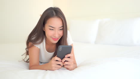 Junge-Lächelnde-Schöne-Asiatin,-Die-Auf-Dem-Bett-Liegt,-Chattet-Auf-Dem-Smartphone