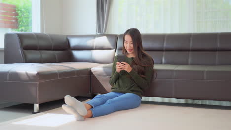 Asiatische-Frau-Sitzt-Auf-Dem-Boden-In-Der-Nähe-Des-Sofas-Und-Schreibt-Auf-Dem-Smartphone