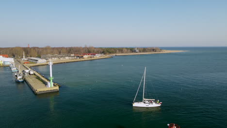 Small-white-sailboat-enters-the-harbor-with-sails-rolled-up,-calm-water-of-Puck-Bay,-Hel,-Poland-aerial