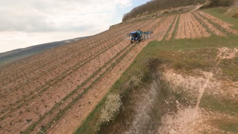 An-FPV-drone's-view-while-orbiting-downwards-towards-a-tractor-sprayer-fertilizing-the-Vineyards-in-Chablis,-France
