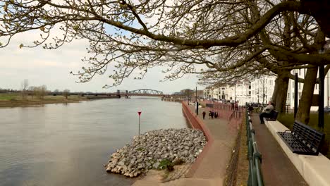 Time-lapse-showing-people-strolling-by-and-relaxing-along-the-river-IJssel-embankment-of-Hanseatic-Dutch-city-Zutphen-with-water-and-cargo-ship-passing-by