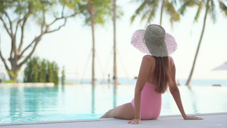 Back-view-of-young-woman-with-pink-swimsuit-and-large-hat-sitting-on-edge-of-resort-pool