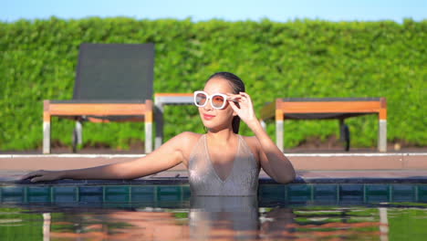 Lächelnde-Asiatische-Frau-Mit-Weißem-Badeanzug-Und-Vintage-Sonnenbrille,-Die-Sich-Im-Poolwasser-Entspannt