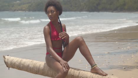 Erstaunliches-Afrikanisches-Model-In-Einem-Bikini,-Das-Auf-Einem-Baumstamm-In-Den-Sandwellen-Des-Ozeans-Sitzt,-Die-An-Der-Küste-Zusammenbrechen